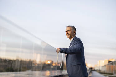 Smiling mature businessman standing by glass railing at sunset - JCCMF09470