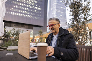 Smiling mature businessman working on laptop at table - JCCMF09473