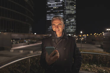 Lächelnder reifer Geschäftsmann, der nachts ein Smartphone benutzt - JCCMF09476