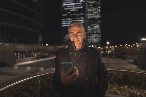 Smiling mature businessman using smart phone at night - JCCMF09476