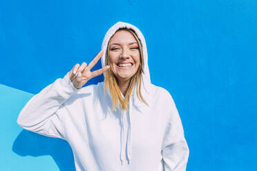 Happy woman making peace sign in front of blue wall - EGHF00721