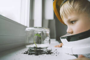 Boy wearing space helmet looking at plant in glass jar - NDEF00358