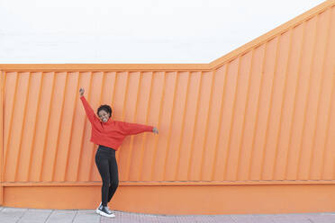 Happy woman with hand raised standing in front of orange wall - LMCF00181