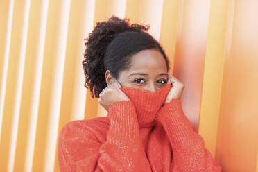 Woman covering face with turtleneck sweater by orange wall - LMCF00182