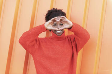Frau, die vor einer orangefarbenen Wand eine herzförmige Geste macht - LMCF00184
