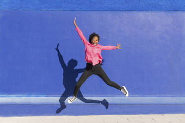 Carefree woman jumping on footpath in in front of blue wall - LMCF00191