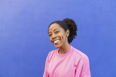 Happy woman with Afro hairstyle in front of blue wall - LMCF00200