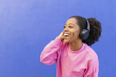 Happy woman listening to music through headphones in front of blue wall - LMCF00202