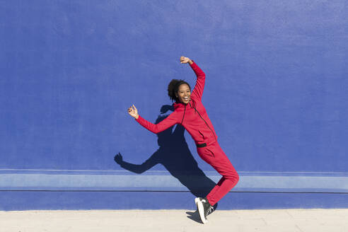 Happy woman balancing on tiptoes in front of blue wall - LMCF00218