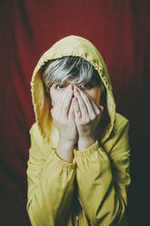 Woman wearing yellow hooded jacket covering mouth against red backdrop - SVCF00365