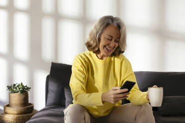 Happy woman using smart phone on sofa at home - EBSF02782