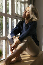 Thoughtful mature woman sitting on window sill at home - EBSF02804