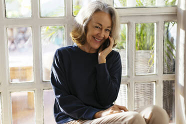 Glückliche reife Frau, die vor einem Fenster mit ihrem Smartphone telefoniert - EBSF02810