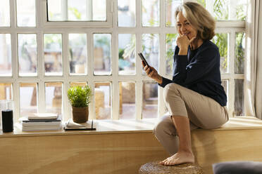 Glückliche Frau mit Smartphone, die zu Hause vor dem Fenster sitzt - EBSF02817