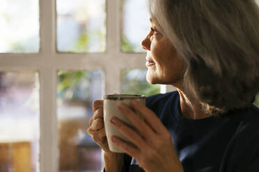 Lächelnde Frau mit Kaffeetasse schaut durch ein Fenster - EBSF02821