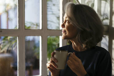 Smiling mature woman with coffee cup looking through window - EBSF02822