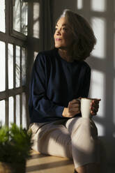 Nachdenkliche Frau mit Kaffeetasse schaut durch das Fenster - EBSF02823