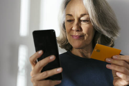 Ältere Frau mit Kreditkarte und Smartphone vor einer Wand - EBSF02832