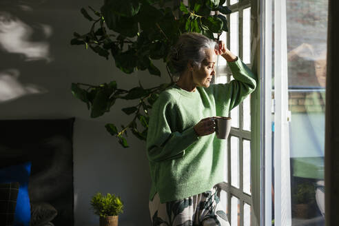 Nachdenkliche Frau lehnt mit Kaffeetasse am Fenster - EBSF02847