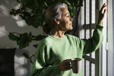 Nachdenkliche Frau mit Kaffeetasse, die zu Hause am Fenster steht - EBSF02848