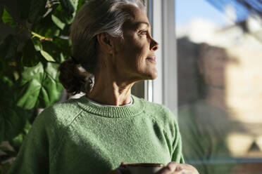 Kontemplative Frau, die durch das Fenster ihres Hauses schaut - EBSF02849