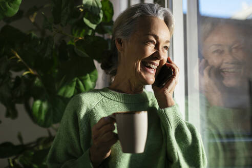 Glückliche reife Frau, die am Fenster zu Hause mit ihrem Mobiltelefon spricht - EBSF02850