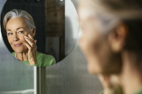 Mature woman applying moisturizer on face looking in mirror - EBSF02853