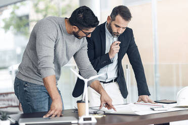 Architekten diskutieren über einen Bauplan in einem Büro mit einem Windkraftanlagenmodell auf dem Schreibtisch - JSRF02421