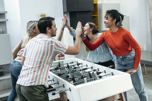 Cheerful colleagues playing foosball and giving high-five at office - JSRF02452