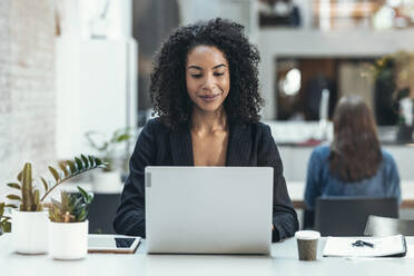 Lächelnde Geschäftsfrau mit Laptop am Schreibtisch am Arbeitsplatz - JSRF02458