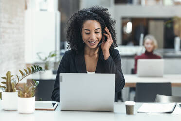 Glückliche Geschäftsfrau, die über ein Headset spricht und einen Laptop im Büro benutzt - JSRF02459