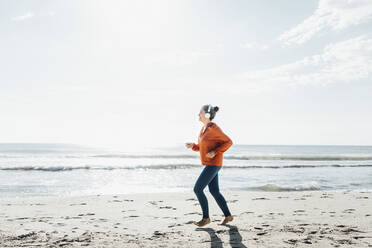 Ältere Frau mit Kopfhörern joggt am Strand - AAZF00080