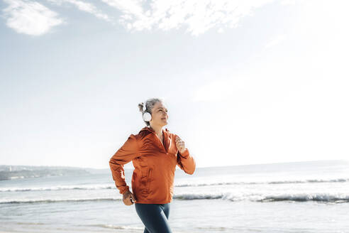 Smiling woman wearing headphones jogging at beach - AAZF00081