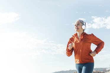 Ältere Frau hört Musik über Kopfhörer und joggt an einem sonnigen Tag - AAZF00082