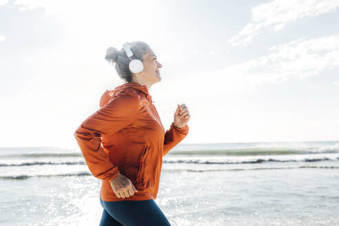 Woman wearing headphones jogging by sea on sunny day - AAZF00085