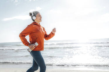 Lächelnde reife Frau mit Wasserflasche joggt am Strand an einem sonnigen Tag - AAZF00088