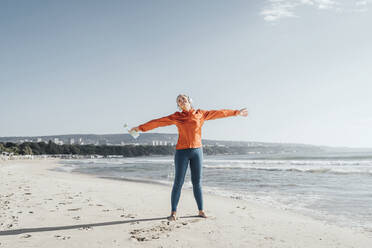 Mature woman standing with arms outstretched on beach - AAZF00089