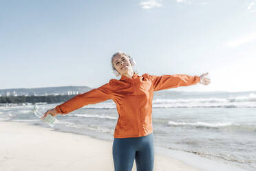 Smiling mature woman standing with arms outstretched on beach - AAZF00090