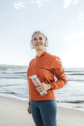 Nachdenkliche reife Frau steht mit Wasserflasche am Strand - AAZF00091