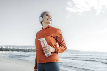 Lächelnde reife Frau mit Wasserflasche am Strand - AAZF00092