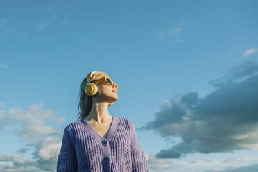 Woman listening to music through wireless headphones under blue sky - VSNF00530