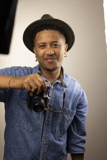 Smiling man wearing hat standing with camera in studio - AXHF00278