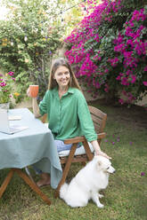 Freelancer drinking coffee sitting with dog in garden - SVKF01282