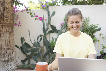 Glücklicher Freiberufler mit Kaffeetasse und Laptop im Garten - SVKF01290