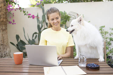 Lächelnder Freiberufler mit Hund am Tisch im Garten - SVKF01291