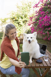 Happy woman taking selfie with dog through smart phone in garden - SVKF01298