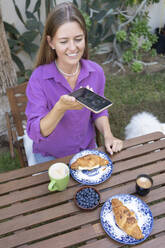 Glückliche Frau, die ein Selfie von Croissants auf einem Tisch im Garten macht - SVKF01306