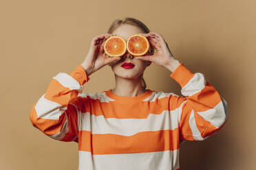 Frau hält Orangenscheiben über die Augen vor braunem Hintergrund - VSNF00534