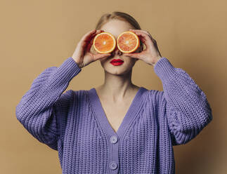 Woman with slices of oranges over eyes against brown background - VSNF00539