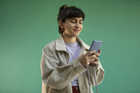 Smiling young woman using mobile phone against green background - AXHF00293